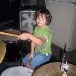 A young boy with curly hair plays a drum set in a room with a fan in the background.