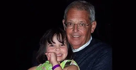 An older man with glasses and a young girl smile against a dark background.