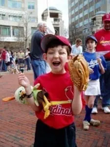 A young boy in a red Cardinals shirt and hat holds a baseball glove and a stuffed toy.