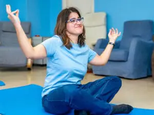 Young woman with glasses sitting cross-legged on a yoga mat with arms raised.