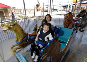 A child in a wheelchair rides a carousel dragon with a woman behind him at an amusement park.