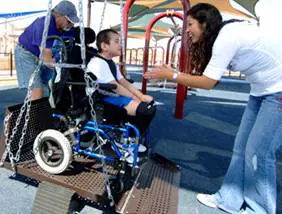 A boy in a wheelchair is on a playground swing with two adults helping him.