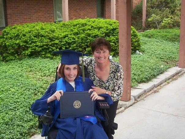 Annie Hopkins in her graduation cap and gown sits in a wheelchair holding her diploma, with a woman smiling behind her.