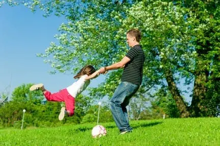 A father and daughter play outside in the grass with a soccer ball.