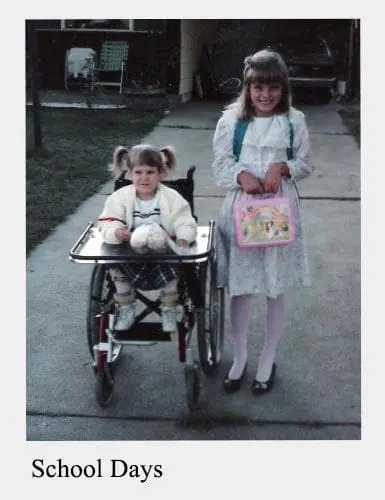 Two children stand on a sidewalk, one in a wheelchair and one holding a lunchbox.