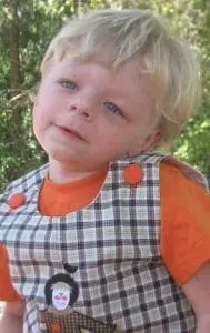 A young child with blonde hair wears an orange shirt and a plaid overall with buttons.