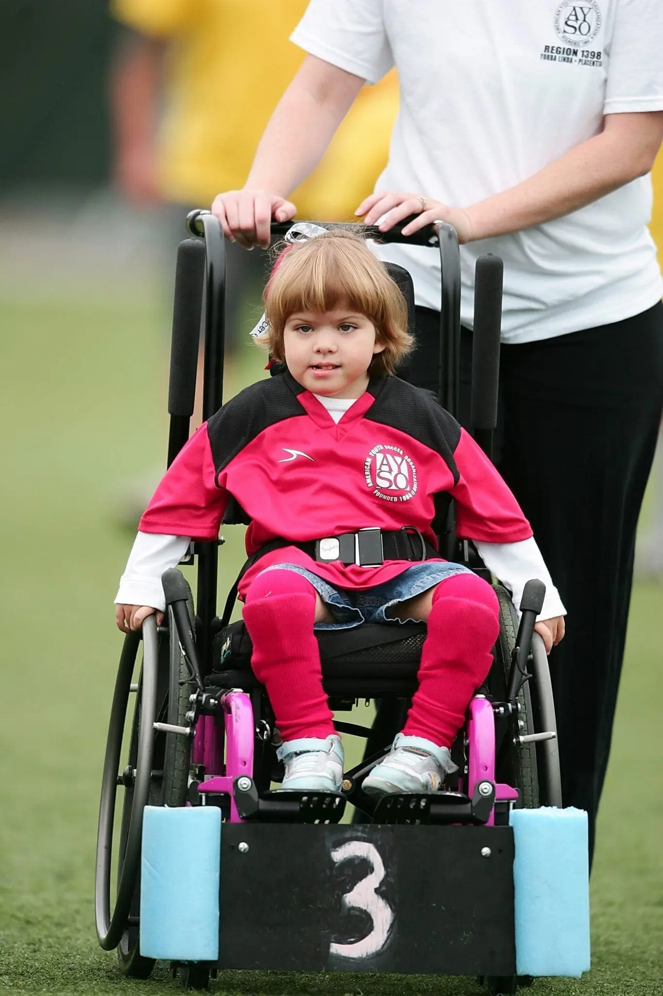 Child in a wheelchair wearing a soccer jersey with a person behind them