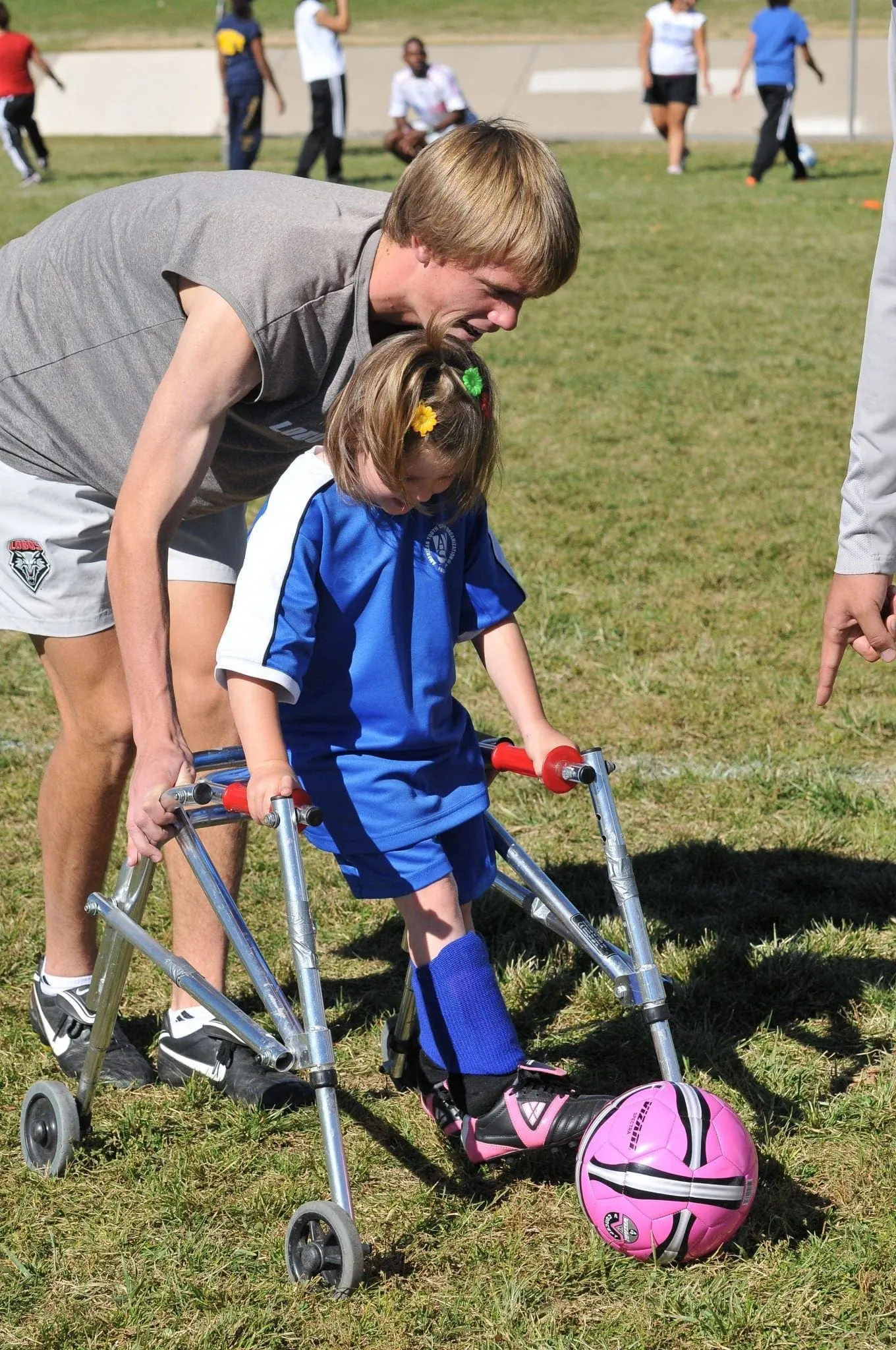 A boy in a blue soccer uniform on a walker reaches for a pink soccer ball with a man behind him.