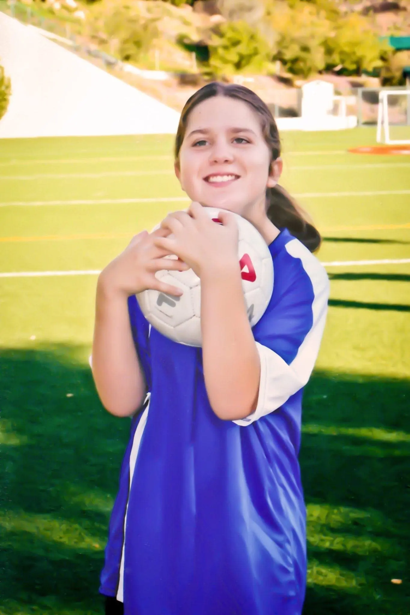 A young girl in a blue and white soccer jersey holds a soccer ball and smiles.