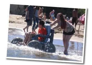 Two people push an adaptive watercraft with a child seated in it into the water on a sandy beach.