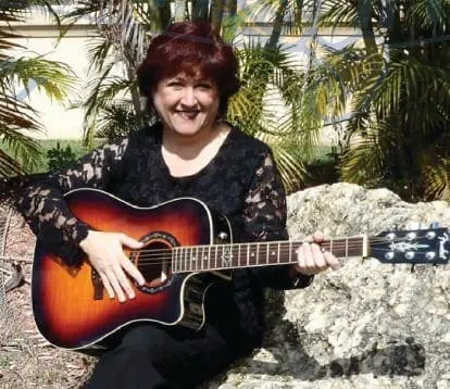 Woman with short, dark, curly hair wearing a black lace top, holding and playing an acoustic guitar outdoors.