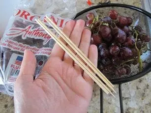 Hand holding wooden skewers next to grapes and a bag of raisins.