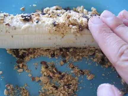 A hand presses crumbled toppings onto a banana half.