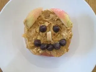 Apple half with peanut butter, chocolate chip face, and apple slice ears on a white plate