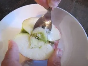 Hands core an apple half with a fork over a white bowl to make a scary face.