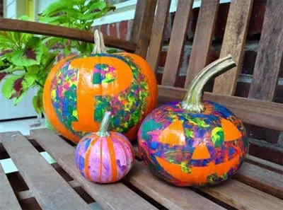 Three pumpkins decorated with colorful paint splatters sit on a wooden bench.