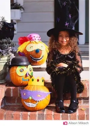 A young girl in a witch costume sits beside decorated pumpkins.