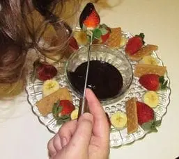 A hand uses a skewer to dip a strawberry into a bowl of chocolate fondue surrounded by fruit and crackers.