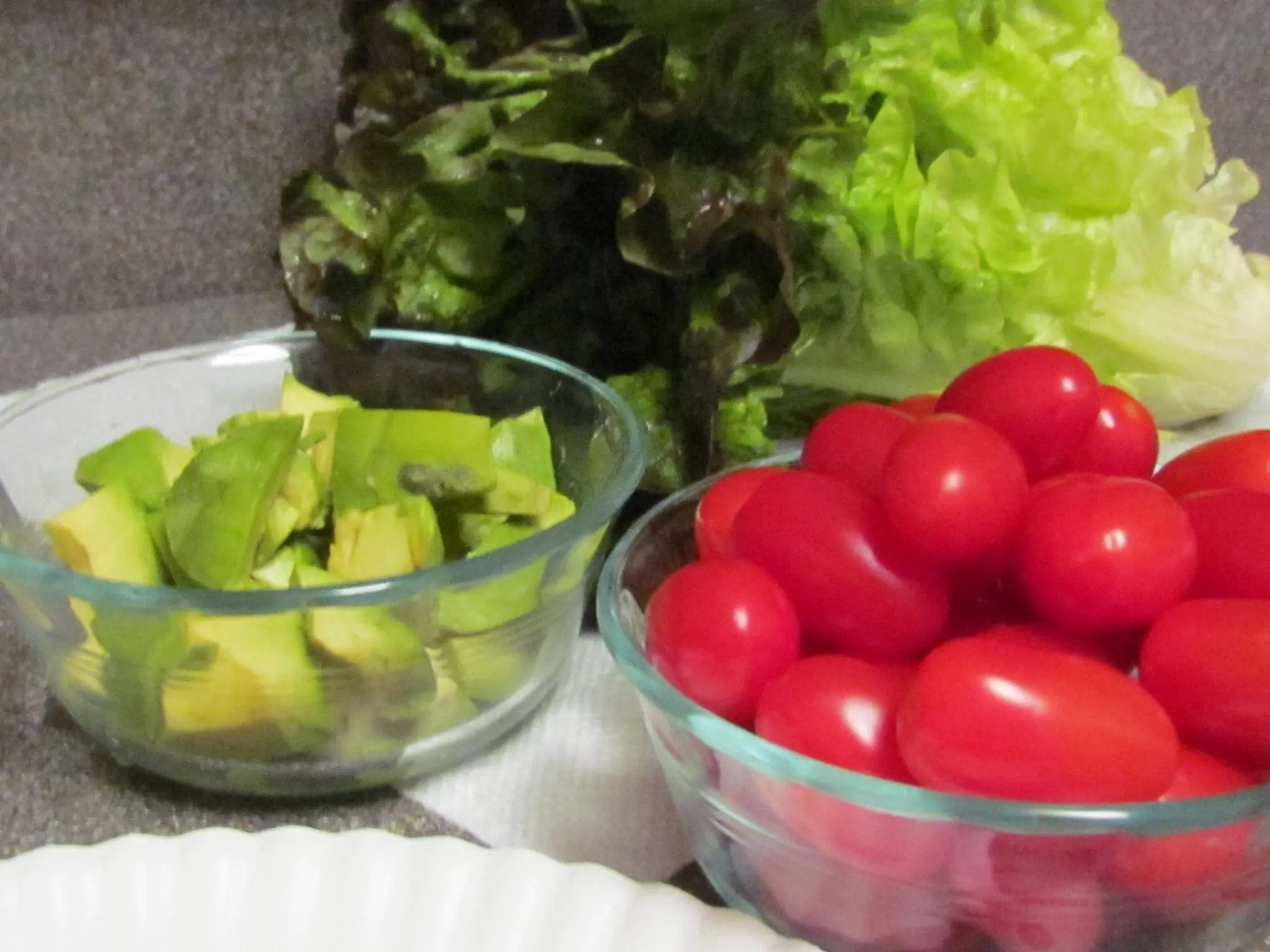 Bowls of chopped avocado and cherry tomatoes next to fresh red leaf lettuce leaves.