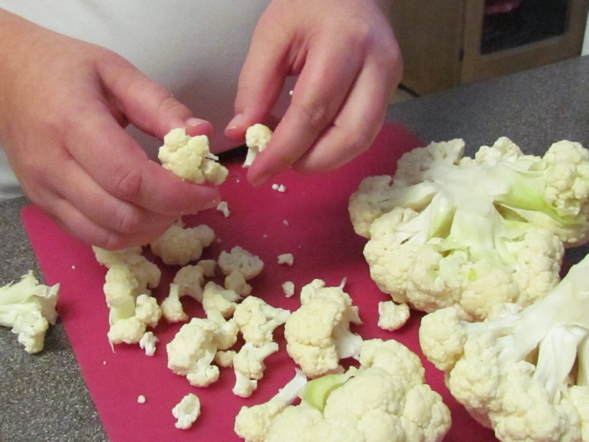 Hands breaking apart cauliflower florets on a pink cutting board.
