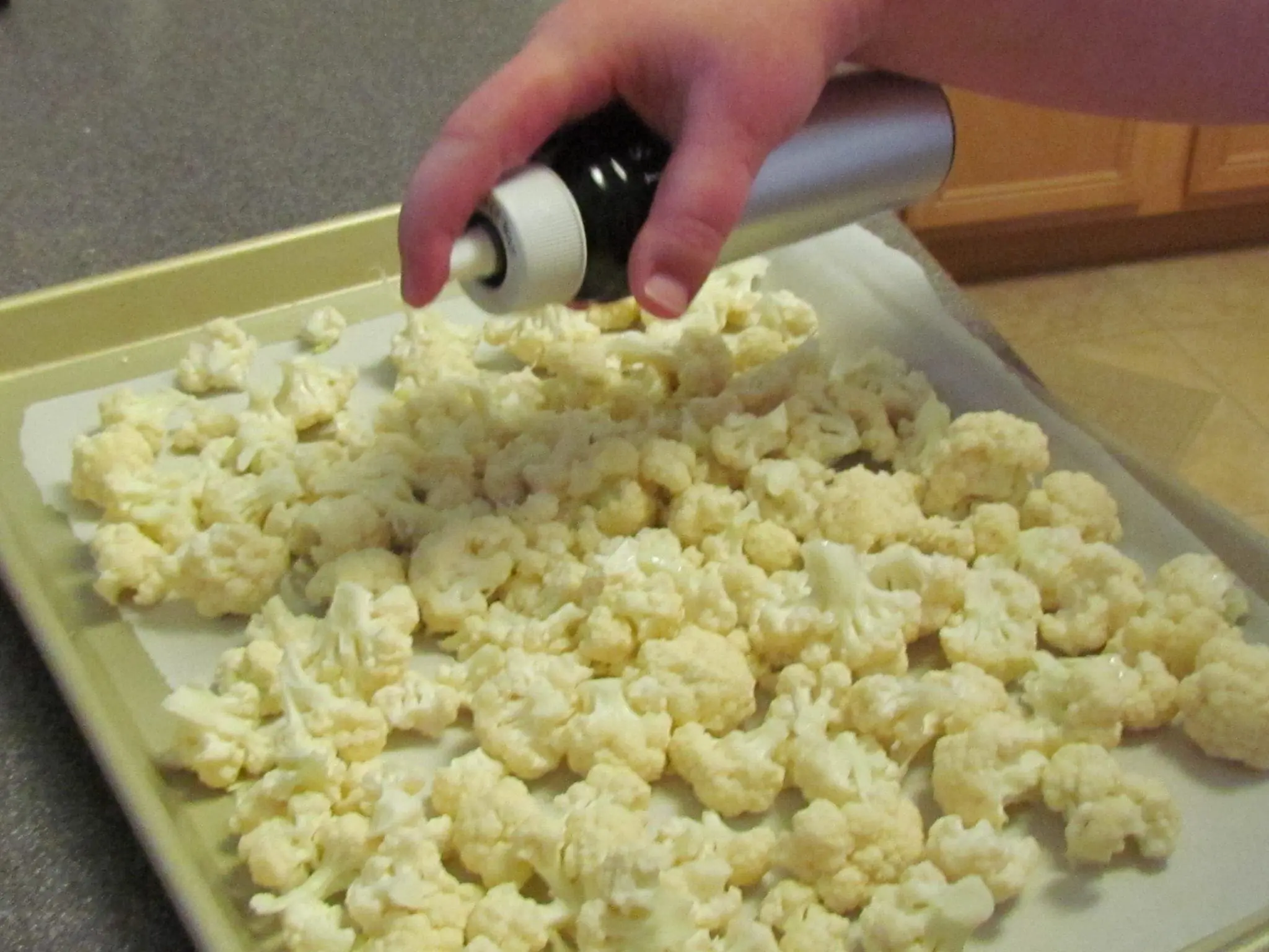 Hand sprays oil onto cauliflower florets on a baking sheet.