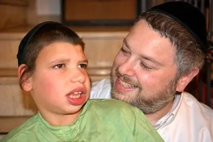 A man with a beard smiles at a boy wearing a black yarmulke and a green shirt.