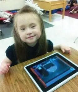 Young girl with Down syndrome smiling at a tablet computer.