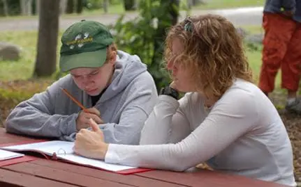 Two people sit at a picnic table with notebooks and pencils.