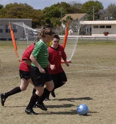 Soccer players of varying abilities run after a ball near a net.