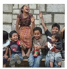 Children laugh and wave while sitting together in front of a brick wall.