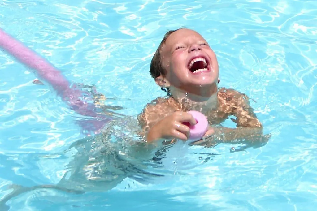 A child laughs while playing in a pool with a pink pool noodle.