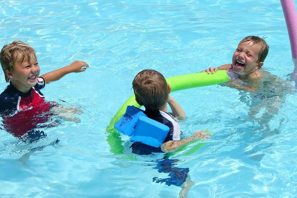 Three children play in a swimming pool with pool noodles.