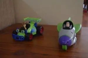 Two Toy Story themed toys, a green race car and a gray and purple airplane, sit on a wooden surface.