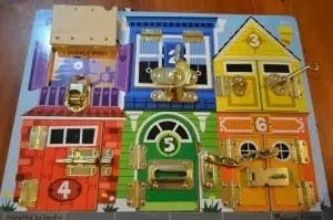 Wooden puzzle board with colorful houses featuring various locks and latches for children to practice fine motor skills.