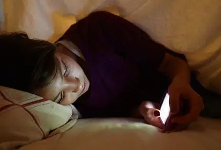 Teenager lying in bed, looking at a glowing smartphone screen in the dark.