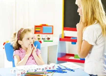 Speech therapist works with a young girl at a table with alphabet blocks and art supplies.