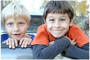 Two young boys lean over a dark surface, one smiling broadly, the other looking forward.