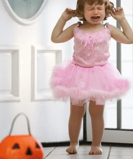 A young child in a pink tutu cries while standing near a jack-o'-lantern candy bucket.