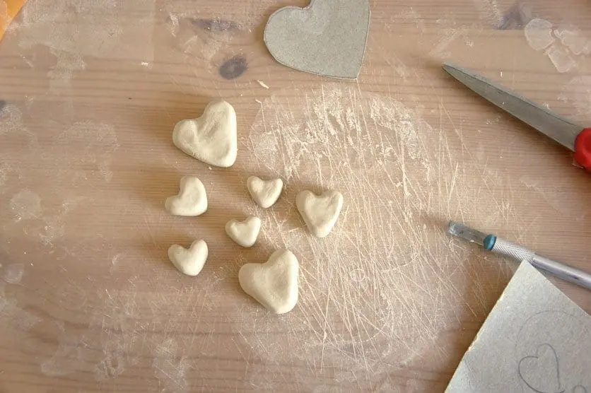 Several clay heart shapes on a wooden surface next to a craft knife and scissors.