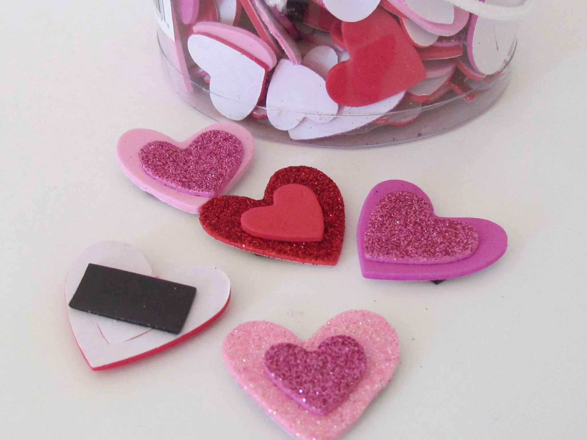 Crafted foam hearts in various shades of pink and red, some with glitter, sit on a white surface near a jar of similar heart shapes.