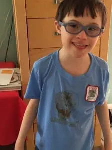 Young boy with Down syndrome and glasses smiles while wearing a blue t-shirt.