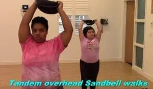 Two people perform tandem overhead sandbell walks.