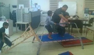 A man assists a child on a trampoline as another child climbs on a ramp.