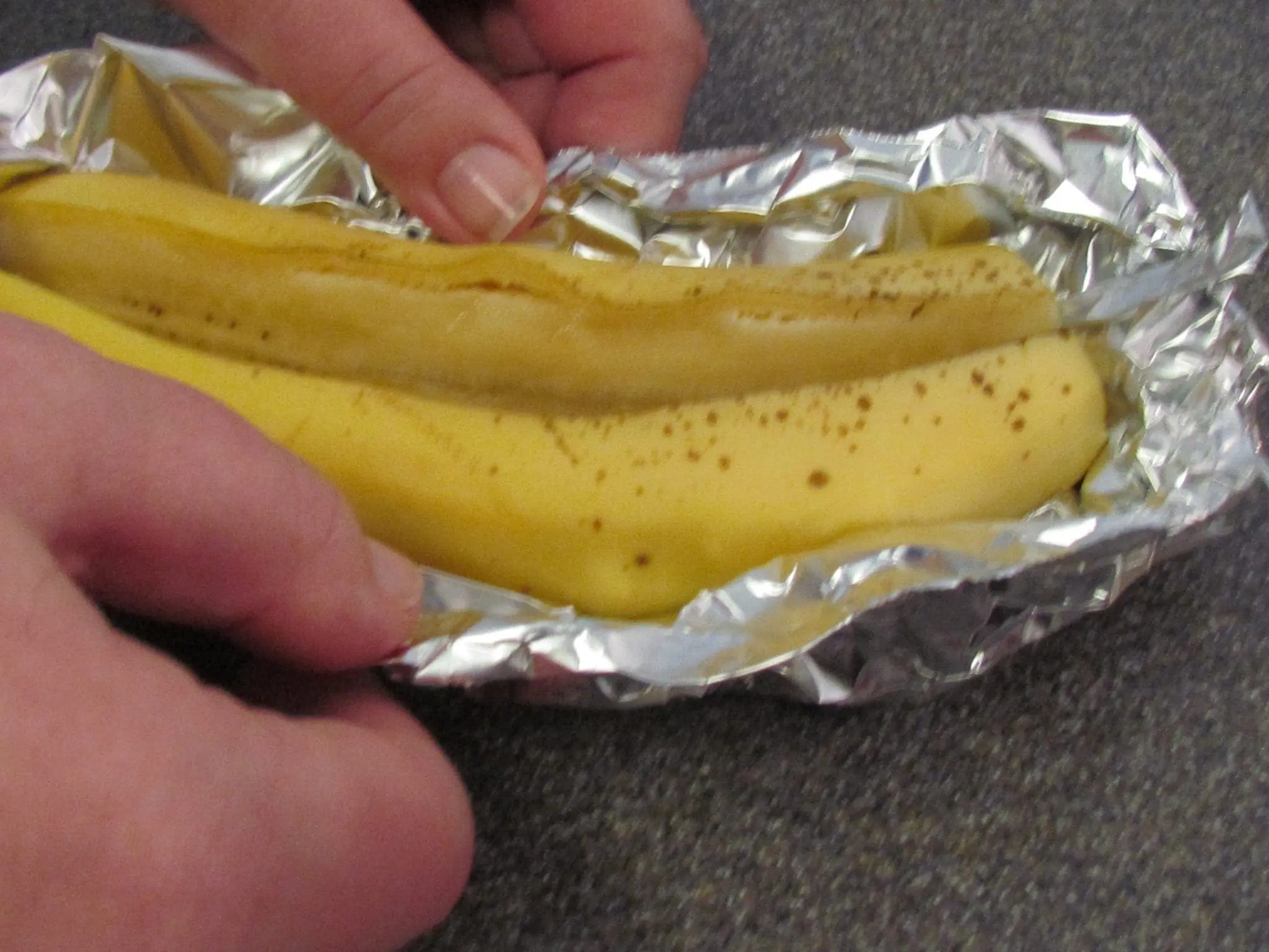 Hands wrap a banana in aluminum foil for campfire cooking.