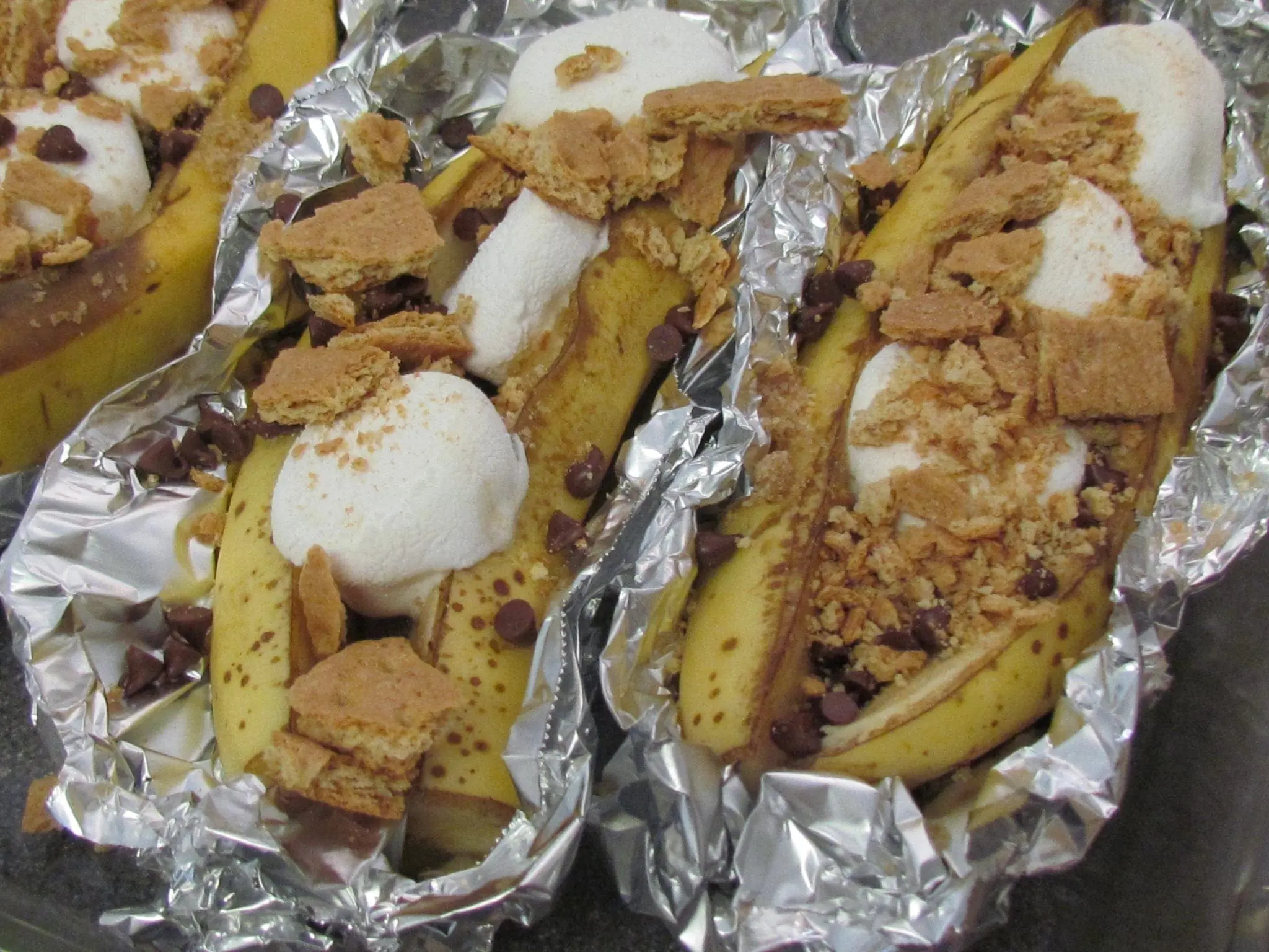 Three banana boats topped with marshmallows and graham crackers in aluminum foil.