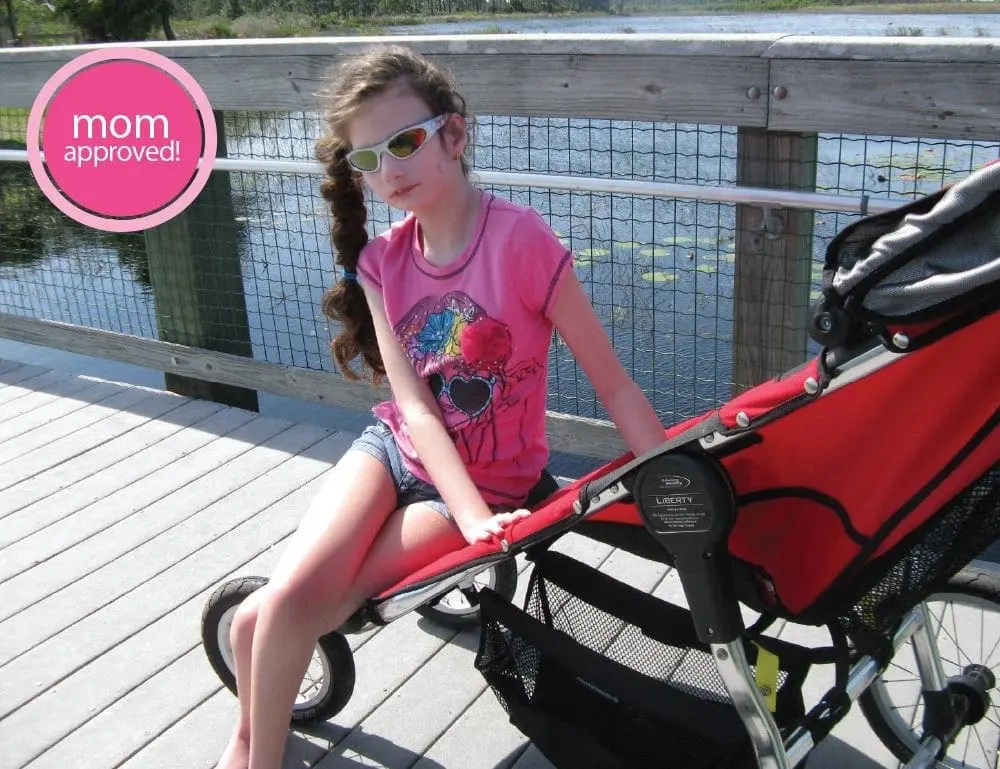 A young girl in sunglasses sits in a stroller on a boardwalk.