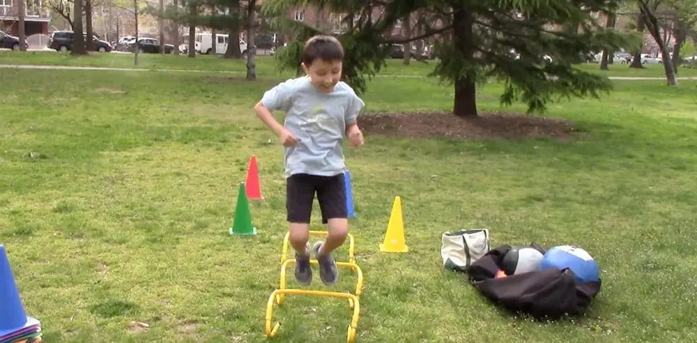 Boy jumping over hurdles on grass with cones and equipment nearby
