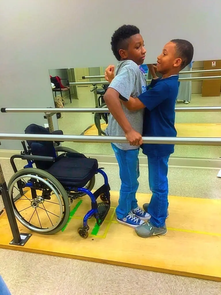 Two young boys hugging next to a wheelchair and parallel bars.