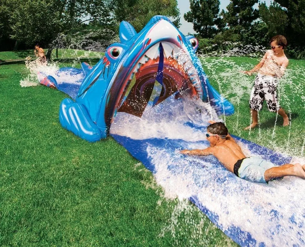 Children slide down a blue inflatable shark water slide on a grassy lawn.