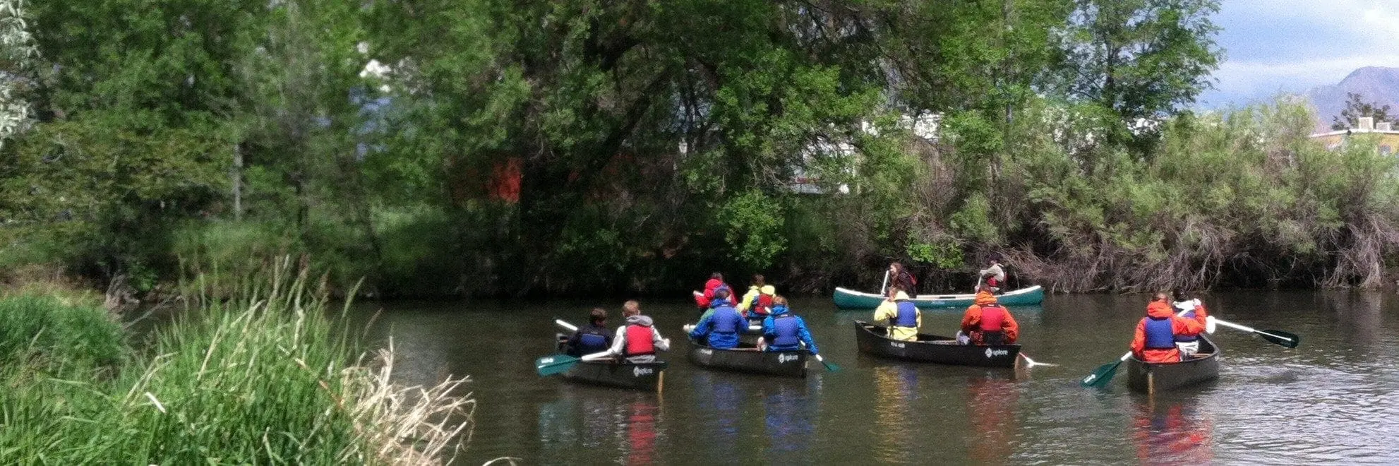 Canoes float down a river lined with trees and tall grass.